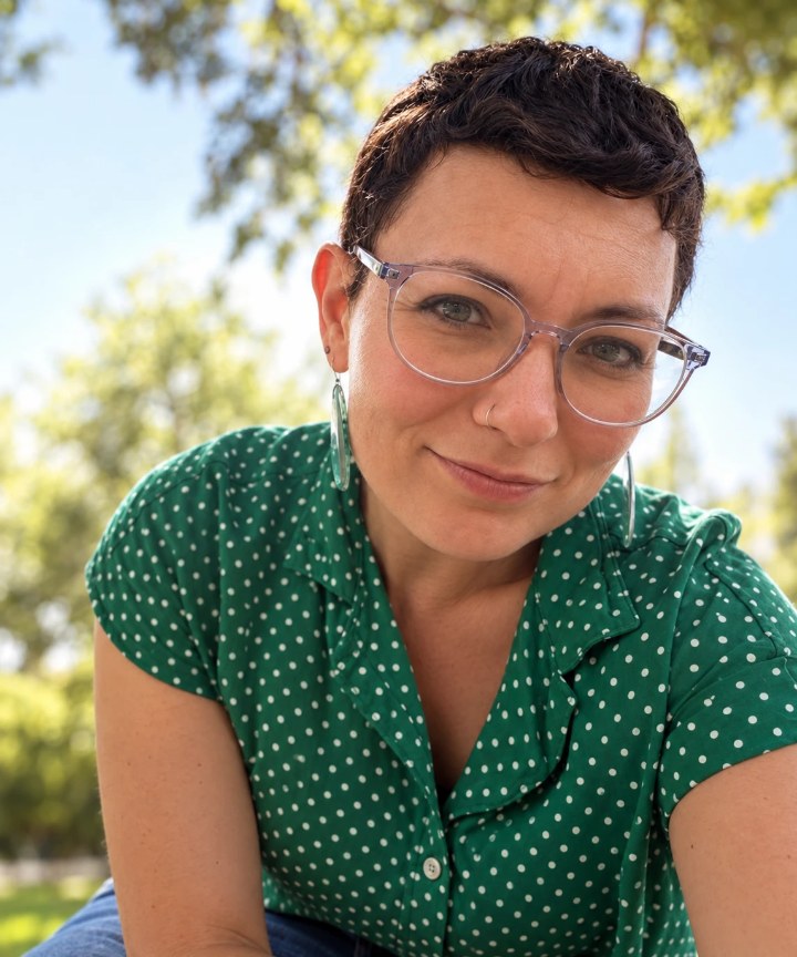 Batami Gold smiling at the camera in glasses and a green polka-dot shirt, outdoors under a tree